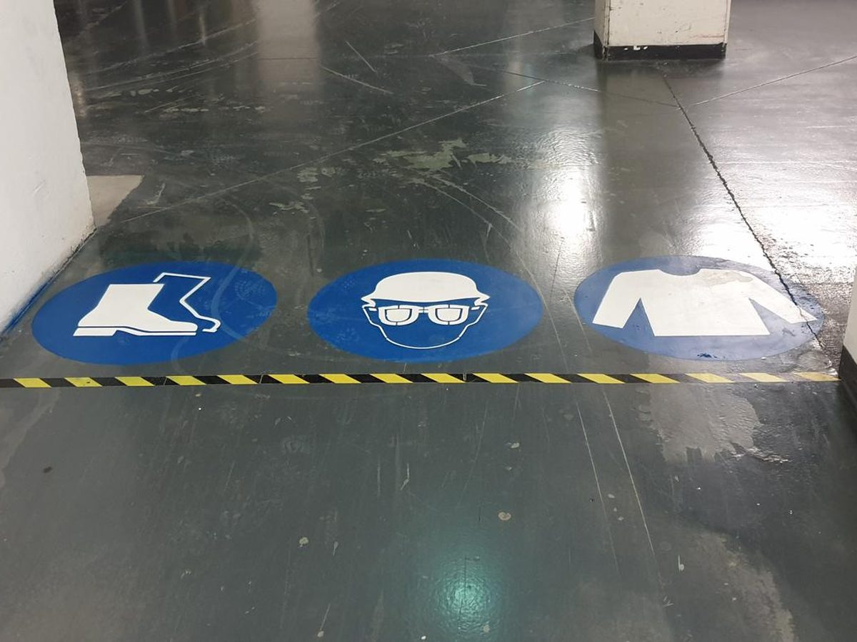 Pictogrammes de sécurité peints au sol : chaussures, lunettes et vêtements de protection, le long d'une bande de délimitation.